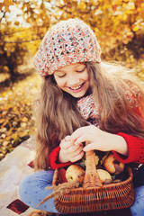happy child girl in knitted scarf and sweater with basket on autumn walk in forest eating apples. Fall harvest, cozy mood.
