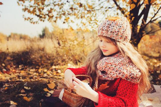 Happy Child Girl In Knitted Scarf And Sweater With Basket On Autumn Walk In Forest Eating Apples. Fall Harvest, Cozy Mood.