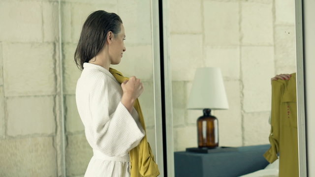 Young, pretty woman in bathrobe trying dresses in front of the mirror in bedroom
