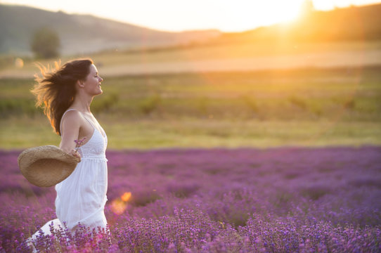 Beautiful Young  Healthy Woman With A White Dress Running Joyfully Through A Lavender Field Holding A Straw Hat Under The Rays Of The Setting Sun.