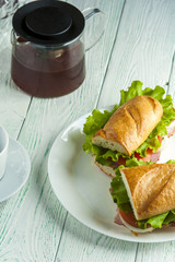 Two tasty sandwiches with ham, vegetable slices an lettuce on white plate, teapot and cup on wooden table. Fast food