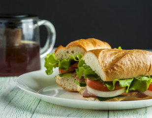 Two sandwiches with baguette, ham, vegetable slices and lettuce on white plate, teapot on wooden table. Fast food. Selective focus.
