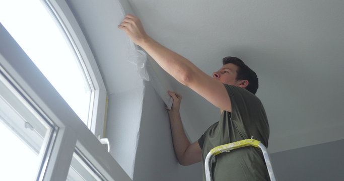 close-up of person standing on a ladder and attaching ribbon tape on wall. window with daylight on the right side of the room.