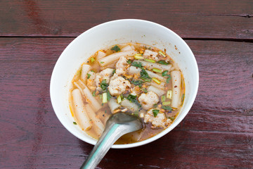A spicy Chinese roll noodle soup with pork and vegetables in a white ceramic/porcelain bowl on dark brown wooden table top