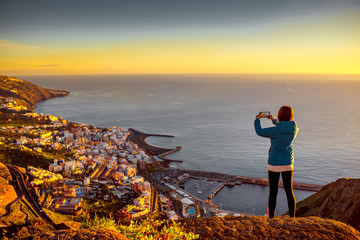 Young female traveler in blue jacket photographing Santa Cruz city standing on the top of the...