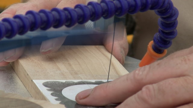 Close up of hands of craftsman holding small piece of flat wood and making fine cut souvenir detail on table top scroll jig using a stencil