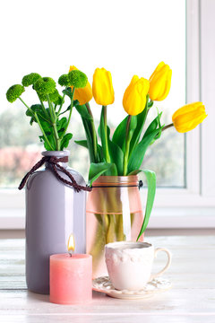 in a vase beautiful and fragrant yellow tulips with green chrysanthemums on a white wooden table and candle with a Cup against the window