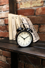 Часы-будильник и книги  стоят на коричневой полке на фоне кирпичной стены