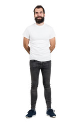 Happy bearded man with hands behind back in white t-shirt and tight jeans looking at camera. Full body length portrait isolated over white studio background. 
