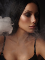 Portrait of the beautiful young girl with dark straight hair, almond-shaped eyes and bronzed perfect covered skin in the black seamless classic bra on the dark background surrounded by the smoke