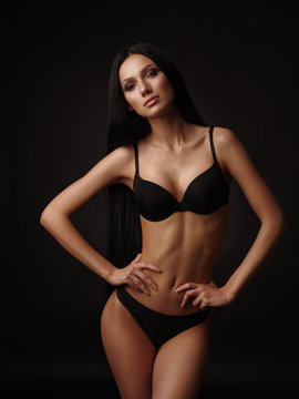 Young beautiful woman with dark straight hair, almond-shaped eyes and tanned skin in the black seamless underwear collection is posing in the studio on the black background