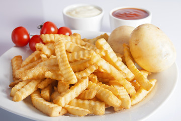Portion of French fries (Crinkle-cut) deep fried, served on a white plate next to white bowls with mayonnaise and ketchup and fresh potato.
