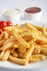 Portion of French fries (Crinkle-cut) deep fried, served on a white plate next to white bowls with mayonnaise and ketchup.