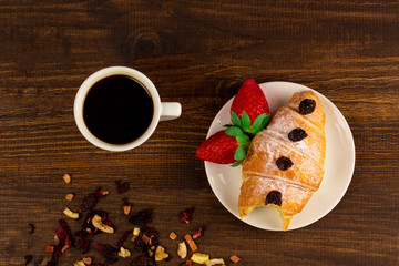 A cup of black tea with dry fruits, croissant with strawberry and wooden table background. Top wiev.