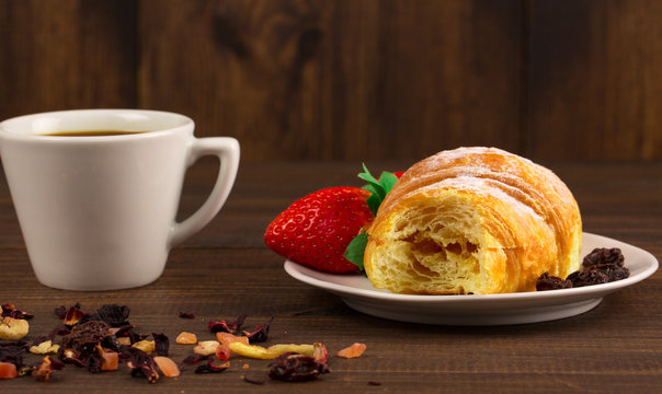 A cup of black tea with dry fruits, croissant with strawberry and wooden table background