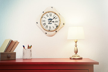 Room interior with red wooden commode, lamp and clock on light wall background