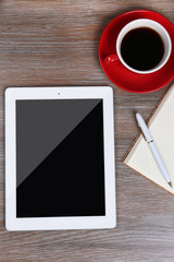 Tablet with cup of coffee and notepad on wooden table