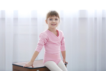 Little cute girl in pink leotard sitting on suitcases at dance studio