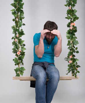 Bearded Man In Jeans, Swinging On Swings And Hamming To The Camera. Wooden Swing Suspended From A Rope Hemp, Rope Wrapped Vine And Ivy. Different Emotions