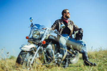 Fototapeta premium Portrait of a young man with beard sitting on his cruiser motorcycle and looking to the sun. Man is wearing leather jacket and blue jeans. Low point of view. Tilt shift lens blur effect