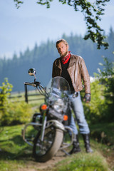 Young handsome biker standing by his custom made cruiser motorcycle on a sunny day with forest on the background. Tilt shift lens blur effect