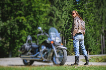 Handsome man with long hair and beard standing near his custom made cruiser motorcycle. Biker is...