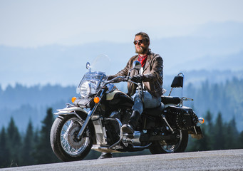 Brutal young man with beard in sunglasses, blue jeans and a leather jacket sitting on the travel motorbike. Sunny day in the mountains