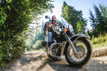 Fototapeta na wymiar Young biker with beard driving his cruiser motorcycle in the forest. Man is wearing leather jacket and blue jeans. Low point of view. Tilt shift lens blur effect