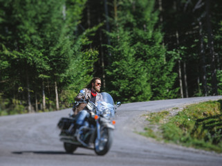 Fototapeta premium Handsome biker with beard driving his cruiser motorcycle by nice road in the forest. Man is wearing leather jacket and blue jeans. Tilt shift lens blur effect