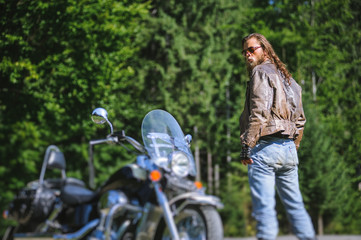 Handsome brutal man with long hair and beard standing near his custom made cruiser motorcycle....