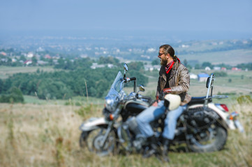 Fototapeta na wymiar biker with beard wearing leather jacket and sunglasses sitting on his motorcycle on a sunny day, holding helmet. Horizontal picture. Tilt shift lens blur effect