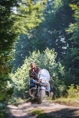 Naklejka premium Young man with beard driving his cruiser motorcycle in the forest and looking aheat on the road. Man is wearing leather jacket and blue jeans. Tilt shift lens blur effect