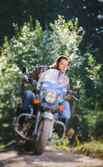 Fototapeta na wymiar biker with beard driving his cruiser motorcycle in the forest. Man is wearing leather jacket and blue jeans. Tilt shift lens blur effect