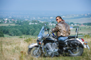Brutal biker with beard wearing leather jacket and sitting on his motorcycle on a sunny day, holding helmet. Horizontal picture. View from the back. Looking to the camera. Tilt shift lens blur effect