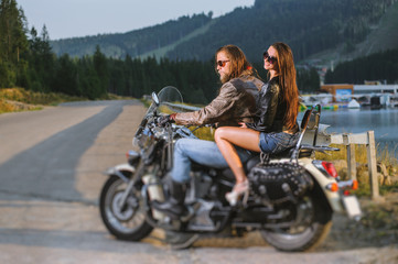 Fototapeta na wymiar Young couple riders sitting together on shiny cruiser motorcycle and starting the travel. Guy and girl wearing leather jackets and sunglasses on a sunny day. Side view. Tilt shift lens blur effect