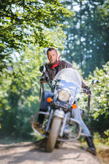 Serious man with beard driving his cruiser motorcycle in the forest. Man is wearing leather jacket and blue jeans. Tilt shift lens blur effect