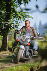 Fototapeta na wymiar Handsome biker with beard driving his cruiser motorcycle in the forest and looking aheat. Man is wearing leather jacket and blue jeans. Tilt shift lens blur effect