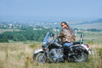 Fototapeta na wymiar Brutal biker with beard wearing leather jacket and sunglasses sitting on his motorcycle on a sunny day. Horizontal picture. View from the back. Looking to the camera. Tilt shift lens blur effect