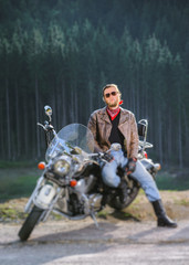 Young handsome biker sitting on his cruiser motorcycle on a sunny day with forest on the background. Man is wearing leather jacket and sunglasses. Vertical photo. Tilt shift lens blur effect