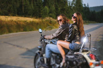 Fototapeta na wymiar Young couple riders sitting together on shiny custom made cruiser motorcycle. Guy and girl wearing leather jackets and sunglasses on a sunny day. Side view. Tilt shift lens blur effect