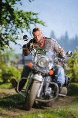 Fototapeta na wymiar Serious biker with beard driving his cruiser motorcycle in the forest. Man is wearing leather jacket and blue jeans. Tilt shift lens blur effect