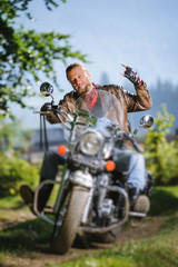 Fototapeta na wymiar Handsome biker with beard driving his cruiser motorcycle in the forest and giving the devil horns gesture and smilling. Man is wearing leather jacket and blue jeans. Tilt shift lens blur effect