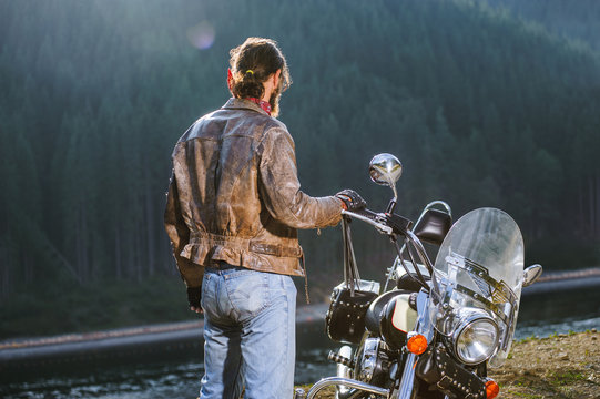 Long Haired Biker Standing Near His Custom Made Cruiser Motorcycle, Wearing Leather Jacket And Blue Jeans. Looking Into Distance. Shot From The Back.