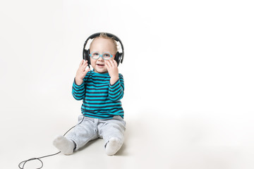 Маленький мальчик в больших наушниках и очках сидит на полу улыбается и пытается снять очки