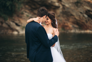 Obraz premium Beautifull wedding couple kissing and embracing near the shore of a mountain river with stones 