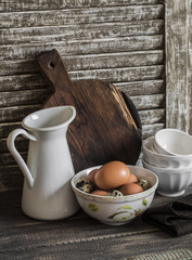 Raw eggs in a bowl, enamelled jug, chopping board and ceramic bowl on a rustic dark wooden background. Kitchen still life in vintage style