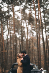 Fototapeta na wymiar Romantic fairytale wedding couple kissing and embracing in pine forest near retro car.