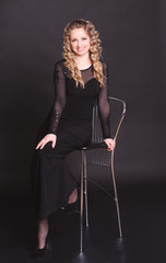 Fototapeta premium Attractive young woman 20-25 year ols sitting on char over black. Smiling blonde girl posing in room, looking at camera. Wearing trendy night dress.