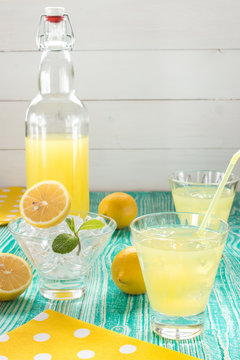lemonade or limonchello in yoke stopper bottle, drink in glasses, sherbet glass with ice cubes decorated by mint leaf, lemon fruits on turquoise colored wooden table with yellow napkin 