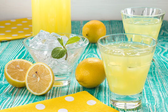 lemonade or limonchello in glasses, sherbet glass with ice cubes decorated by mint leaf, lemon fruits on turquoise colored wooden table with yellow napkin at white polka dots